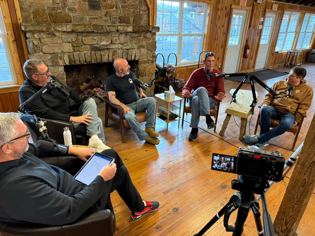 The five friends recording the podcast in a wood cabin, gathered in chairs around microphones by a stone fireplace.